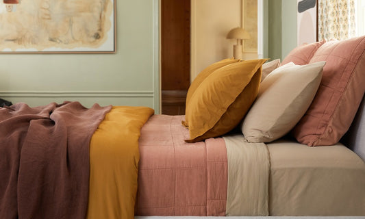 A bed made up with various layers of sheets, blankets, and pillows. Colors are beige, pink, yellow, and brown.
