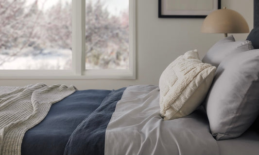 A casually-made bed shows various layers of sheets, blankets, and pillows.