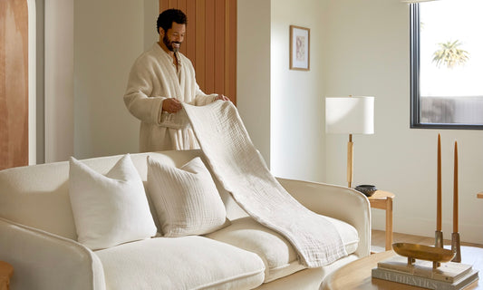 A man wearing a robe folds a towel over a white couch. 